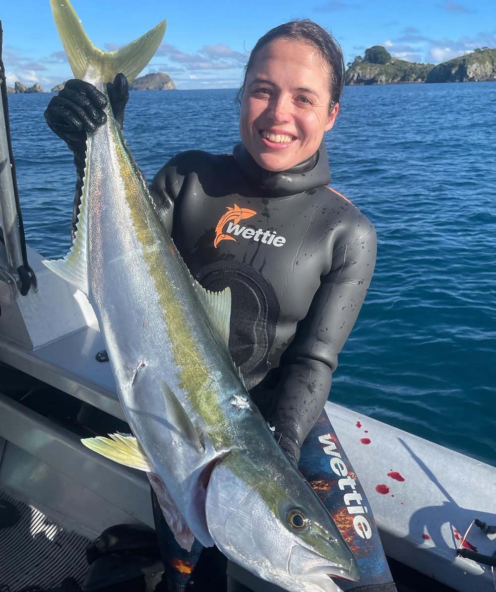 Person in a wetsuit holding a large fish on a boat with a scenic background