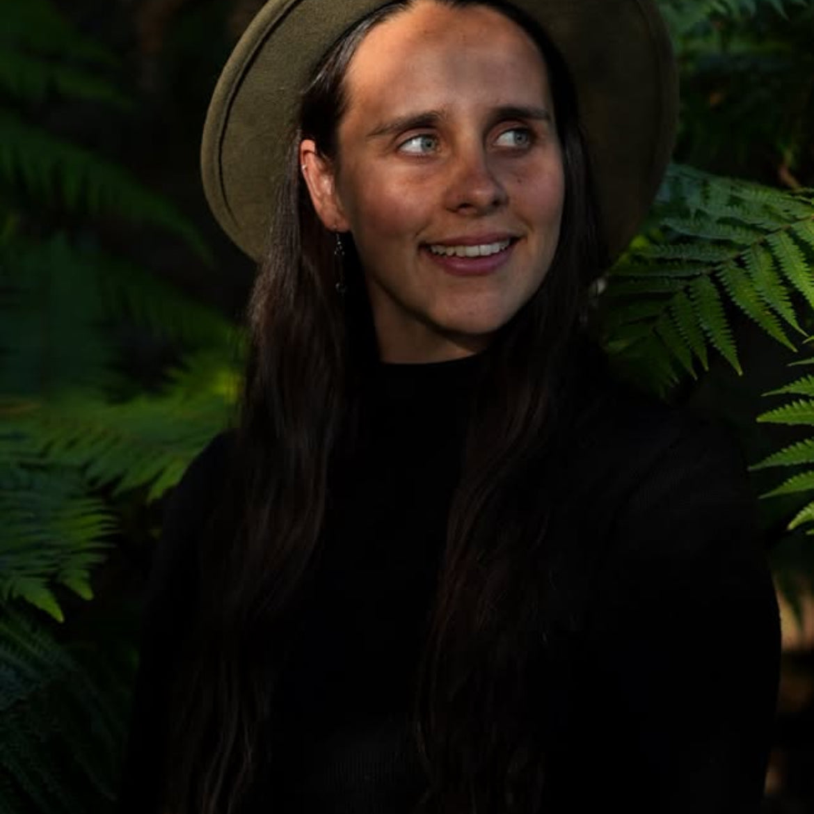 Han wearing a hat and dark clothing in front of green foliage