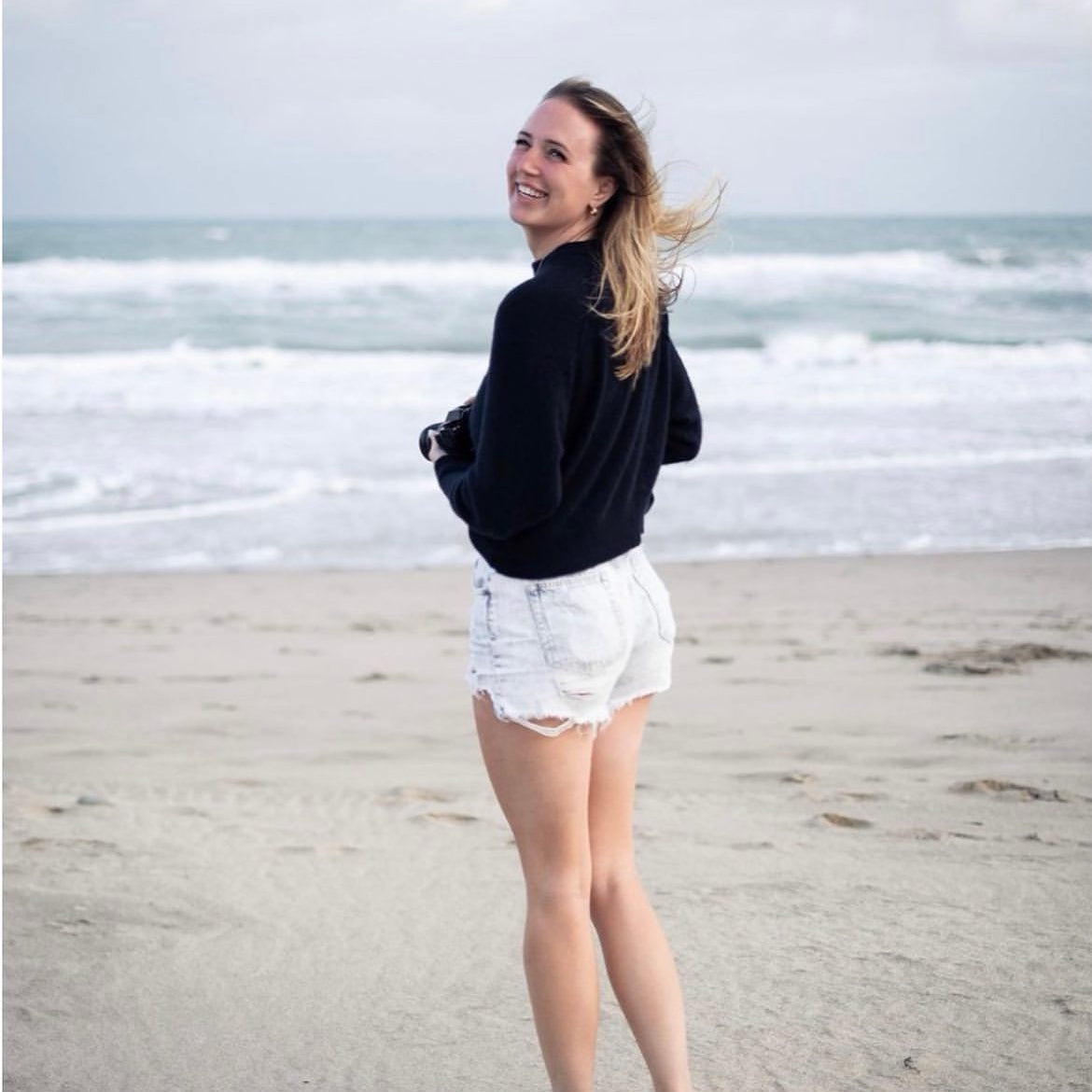 Jenae standing on a beach wearing a black sweater and white shorts.