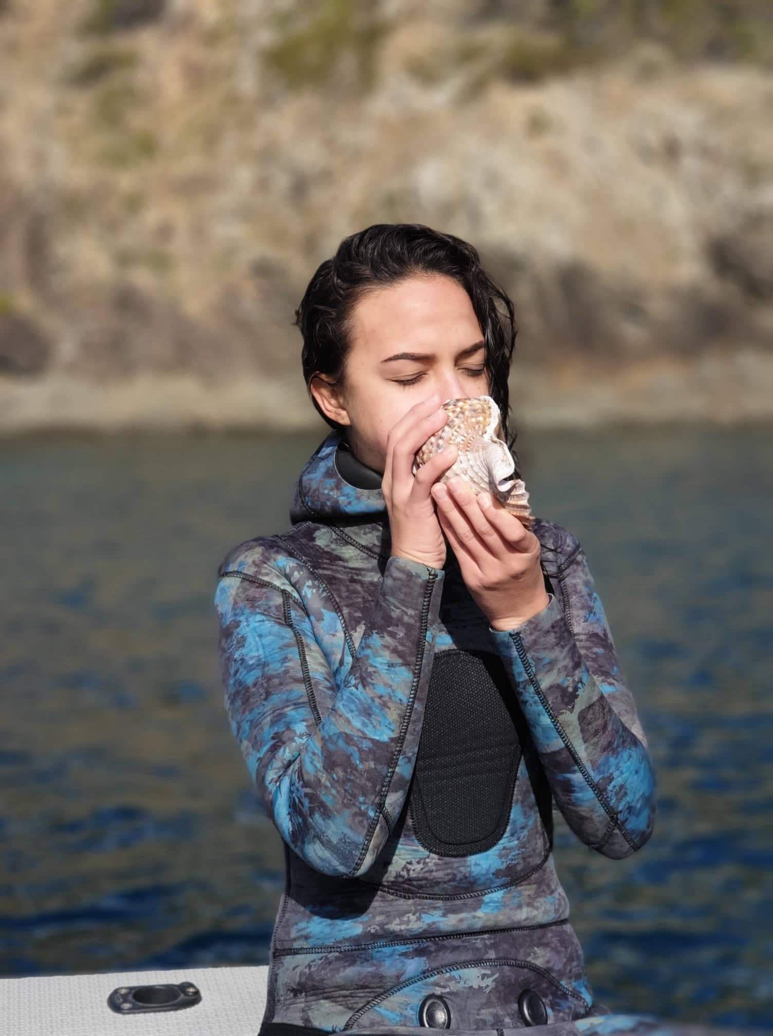 Renee blowing into a shell by a body of water