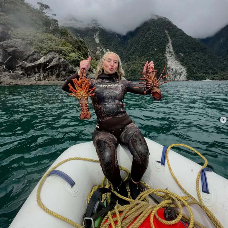 Brooke in a wetsuit holding two lobsters on a boat with a mountainous background