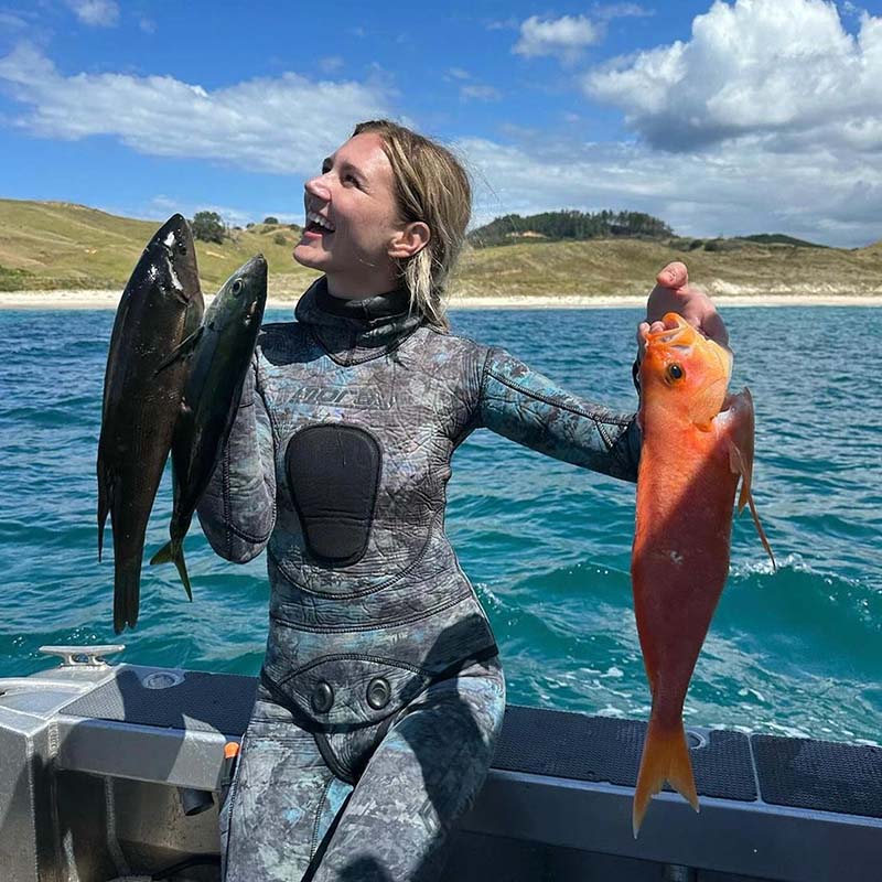 Elena in a wetsuit holding two fish on a boat with a scenic background