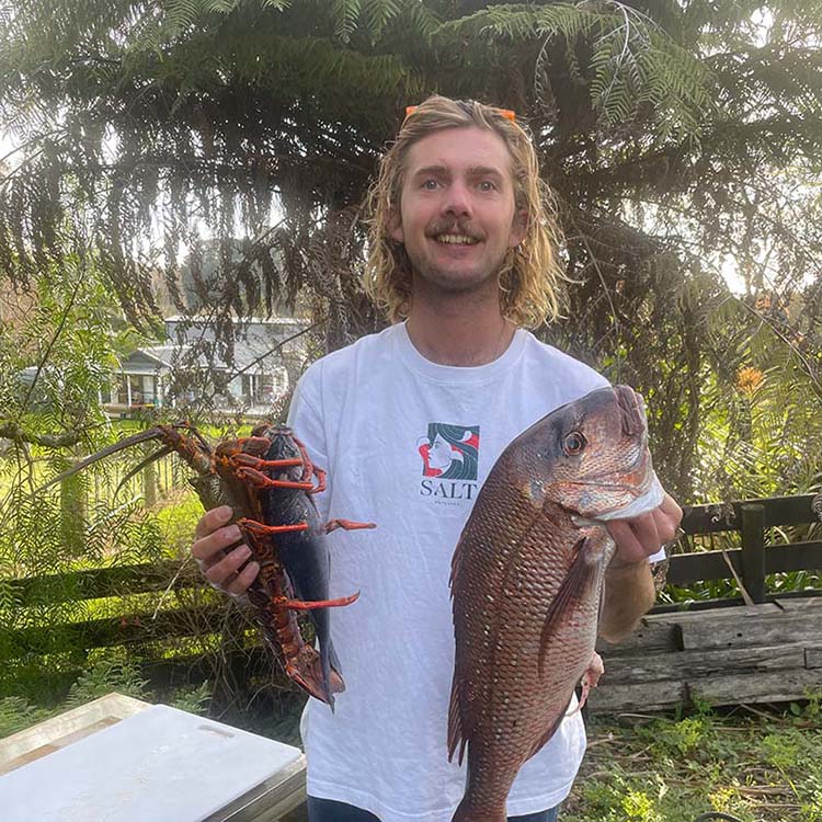 Zac holding a fish and a lobster outdoors with trees and a house in the background